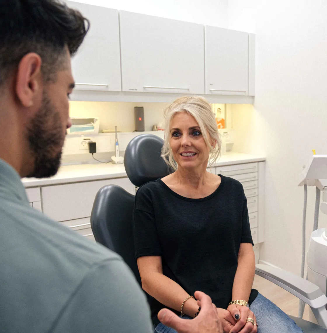 Patient sidder i tandlægestol og lytter til tandlægen der sidder og snakker til hende